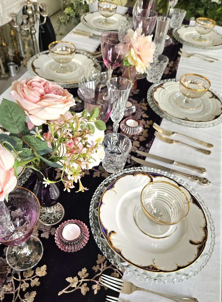 trestle table set up with a deep purple runner embroidered with gold leaves and purple glass and vintage vases filled with pink fake flowers and pink tealight holders the place settings are complete with glass charger plates, white and blue rimmed vintage plates and silver vintage cutlery and cut glass tumblers, wing and champagne flutes for hire