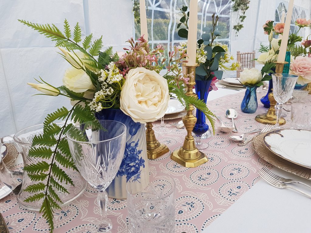 A Garden marquee wedding with vintage blue china vases and jugs filled with wild flowers and blue glass posy vases with faux eucalyptus and roses mixed with brass candlesticks with cream candles  to hire