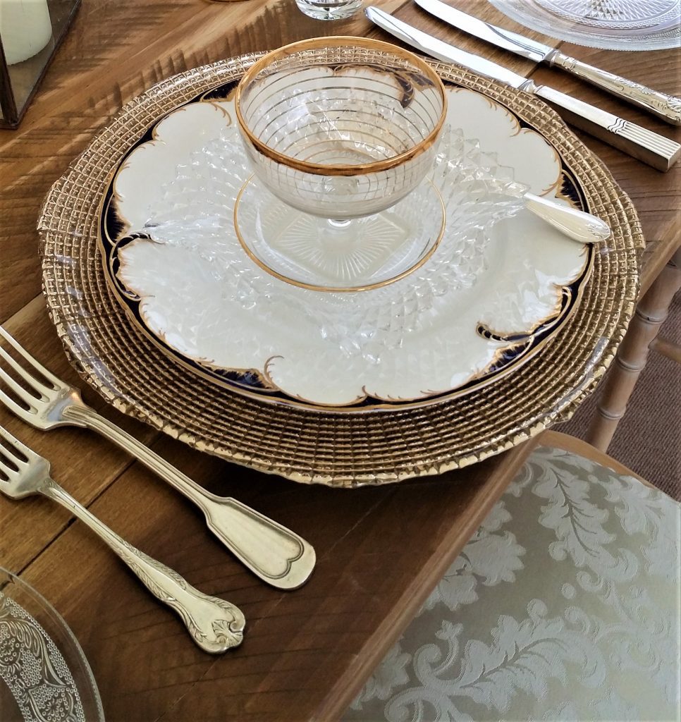 Place setting for wedding or function of a gold charger plate with a white dinner plate with a navy and gold trim on top is a glass posy bowl also with a gold trim, the place setting is finished with a full set of silver vintage cutlery all available to hire