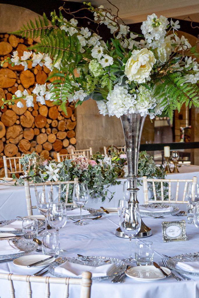 Silver Trumpet vase wedding floral centrepiece with a flower arrangement with ferns, white and pink roses, white and green hydrangeas, gypsophila and white stocks as w round table centrepiece with cut glass charger plates and a classic diner set complete with wine and water glasses and cutlery to hire