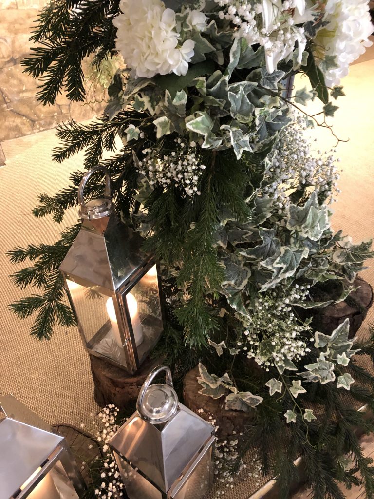 Three silver lanterns on wooden tree stumps in front of a frame of ivy, pine branches and gypsophila, for wedding ideas at great tythe barn, available to hire for weddings and events