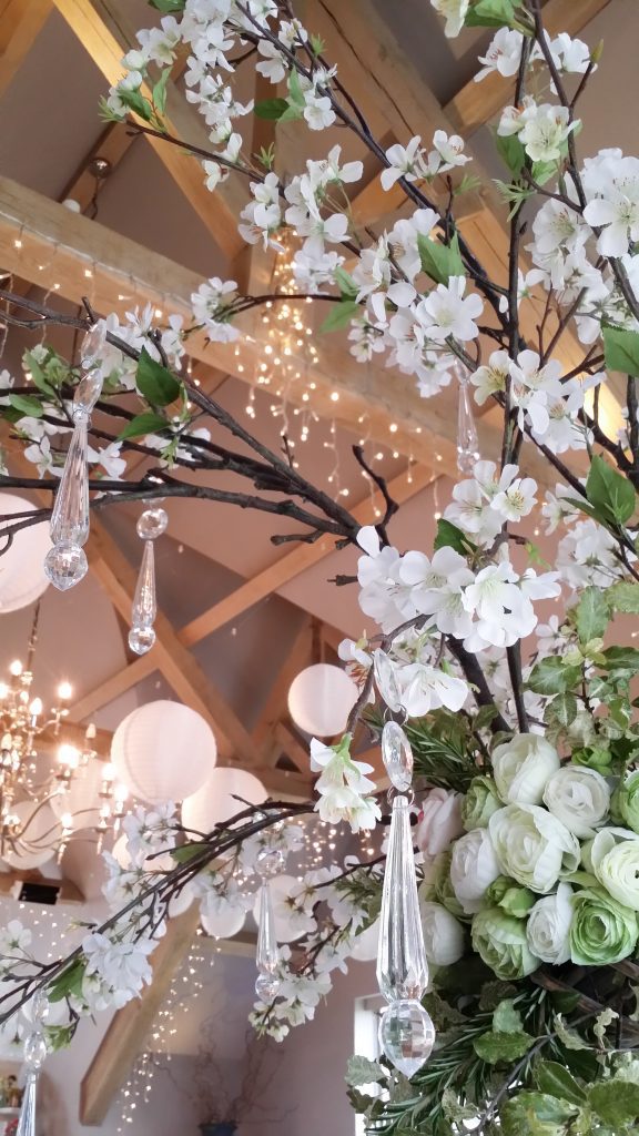 close up of Luxury quality faux flowers in whites and greens, with spring blossom branches of white blossom and clear crystal hanging from them. with a backdrop of fairy lights wrapped around beams in the eaves at Hyde Barn in the Cotswolds