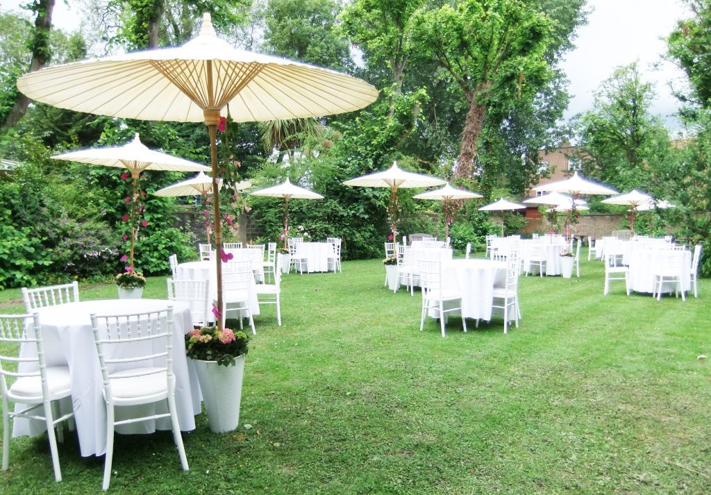 Celebration of life garden set up with 10 small round tables covered in white tablecloths with white bamboo chairs around them. Next to each table stands a white bucket filled with pink hydrangeas and greenery covering the base of a beautiful Japanese style paper and bamboo parasol with strings of Hot pink orchids and baby pink sweet peas hanging rom the spokes