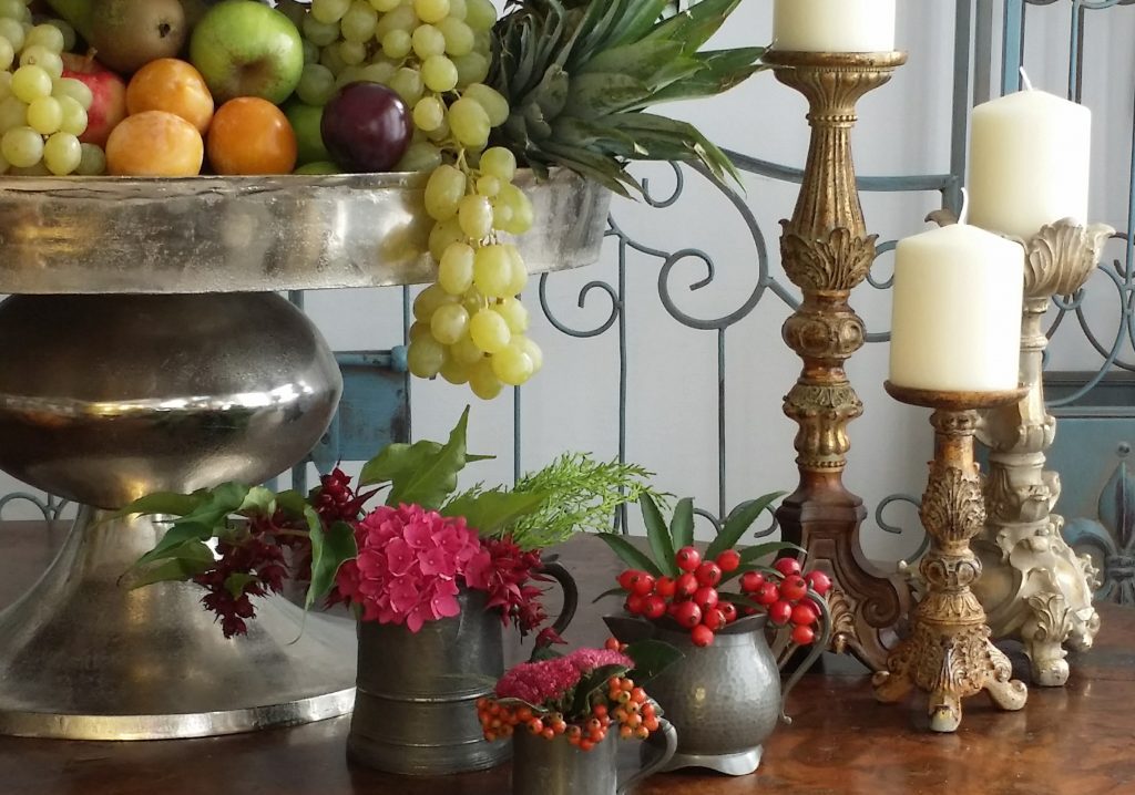 the base of an extra large silver pedestal cake stand with grapes, pineapple plums and pears in it, With vintage beer tankard or pewter mug filled with winter berries and foliage and three gold ornate candlesticks to the right holding church candles