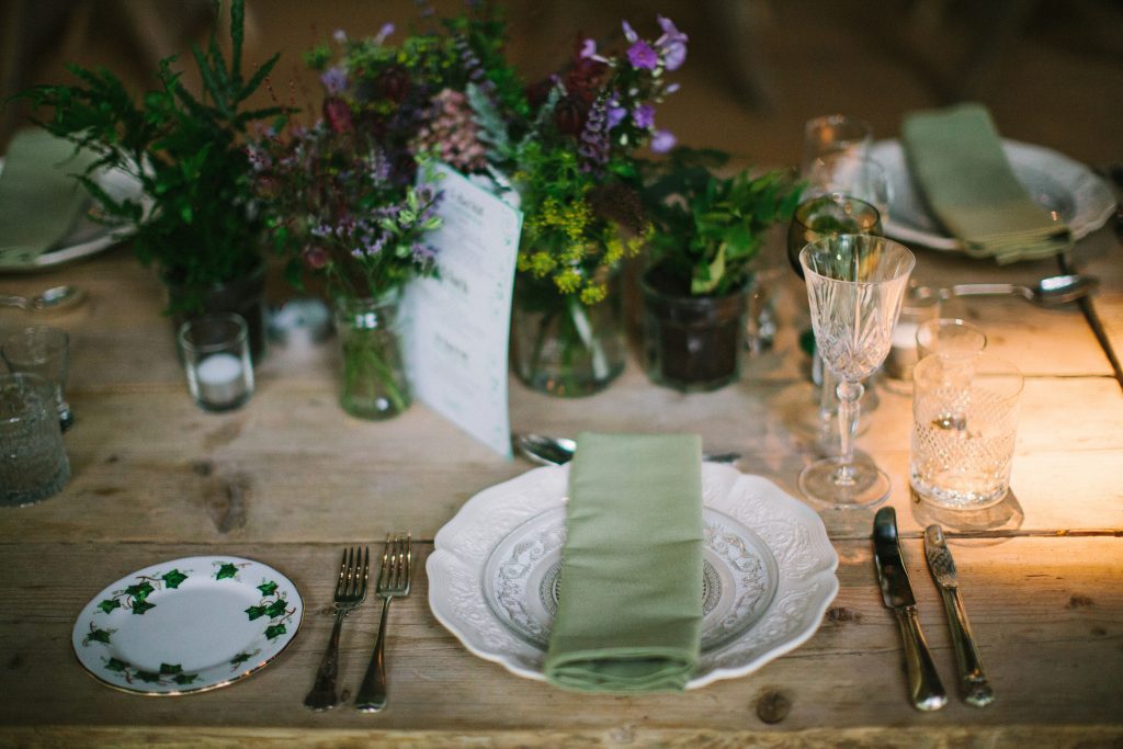 Trestle table set up with the focus on a centrepiece of mixed glass jars filled with wild flowers and a place setting completer with vintage cut crystal wine glasses and water tumbler, mix and match vintage silver cutlery and vintage dinner plate and other crockery and a sage green napkin available to hire for weddings and events